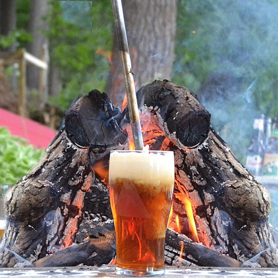 A poker submerged in a glass of beer sitting in front of a campfire